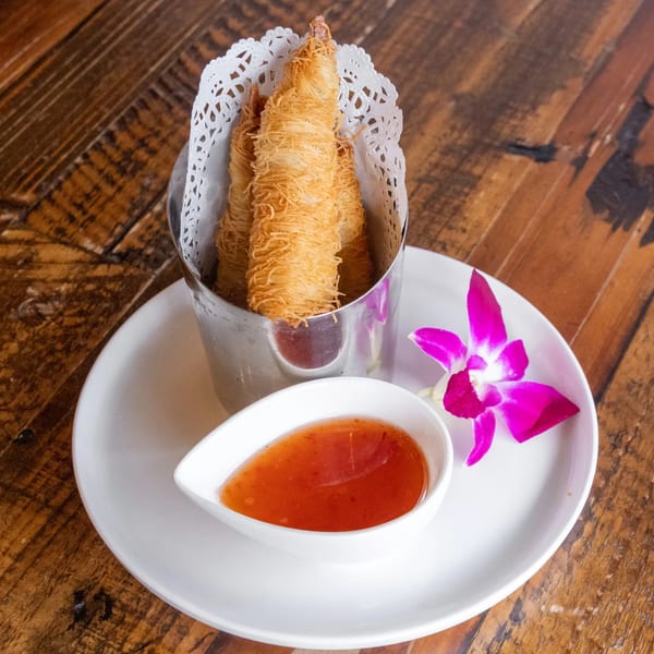 Crispy shrimp rolls in a metal holder with sweet chili sauce and an orchid on a white plate.